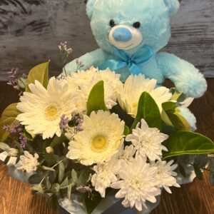 A posy box arrangement of fresh flowers and a teddy bear. Flowers include gerberas and chrysanthemums.