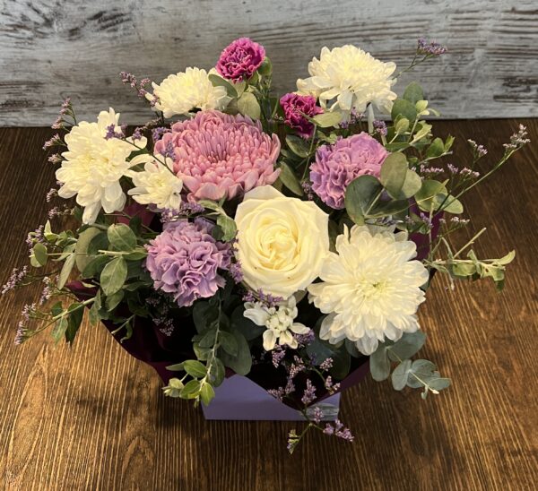 A posy box arrangement in purples, mauves and whites. Flowers include gerberas, roses and disbud chrysanthemum.